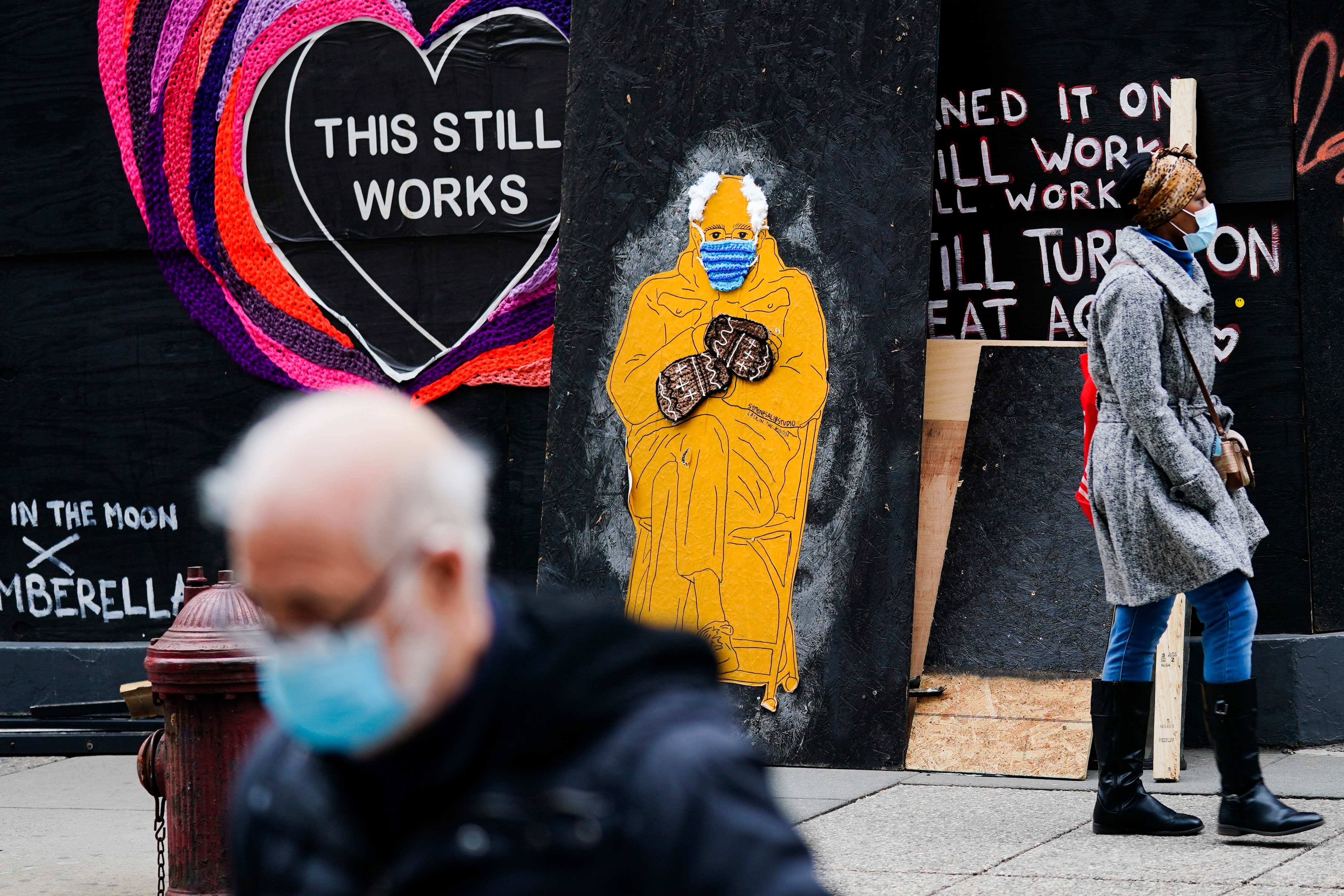 People wearing face masks as a precaution against the coronavirus walk past a depiction of Senator Bernie Sanders. (AP) People wearing face masks as a precaution against the coronavirus walk past a depiction of Senator Bernie Sanders. (AP)