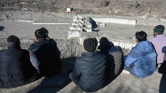 [People sit on a hill overlooking the remains of a dam along a river in Tapovan of Chamoli district on February 8, 2021 destroyed after a flash flood thought to have been caused after glacier burst on February 7. (AFP) - fonte: https://www.hindustantimes.com/india-news/uttarakhand-flash-floods-at-least-26-dead-171-still-missing-amid-rescue-ops-101612828040432.html]