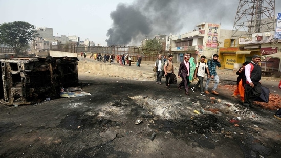 Members of a wedding procession are seen walking on a road littered with debris and damaged vehicles, while smoke rises in the background at Karawal Nagar.(Biplov Bhuyan/ HT Archive)