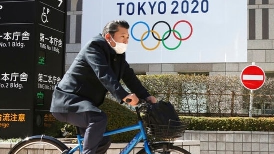 A man wearing a face mask cycles past the logo of the Tokyo Olympics. (AP)
