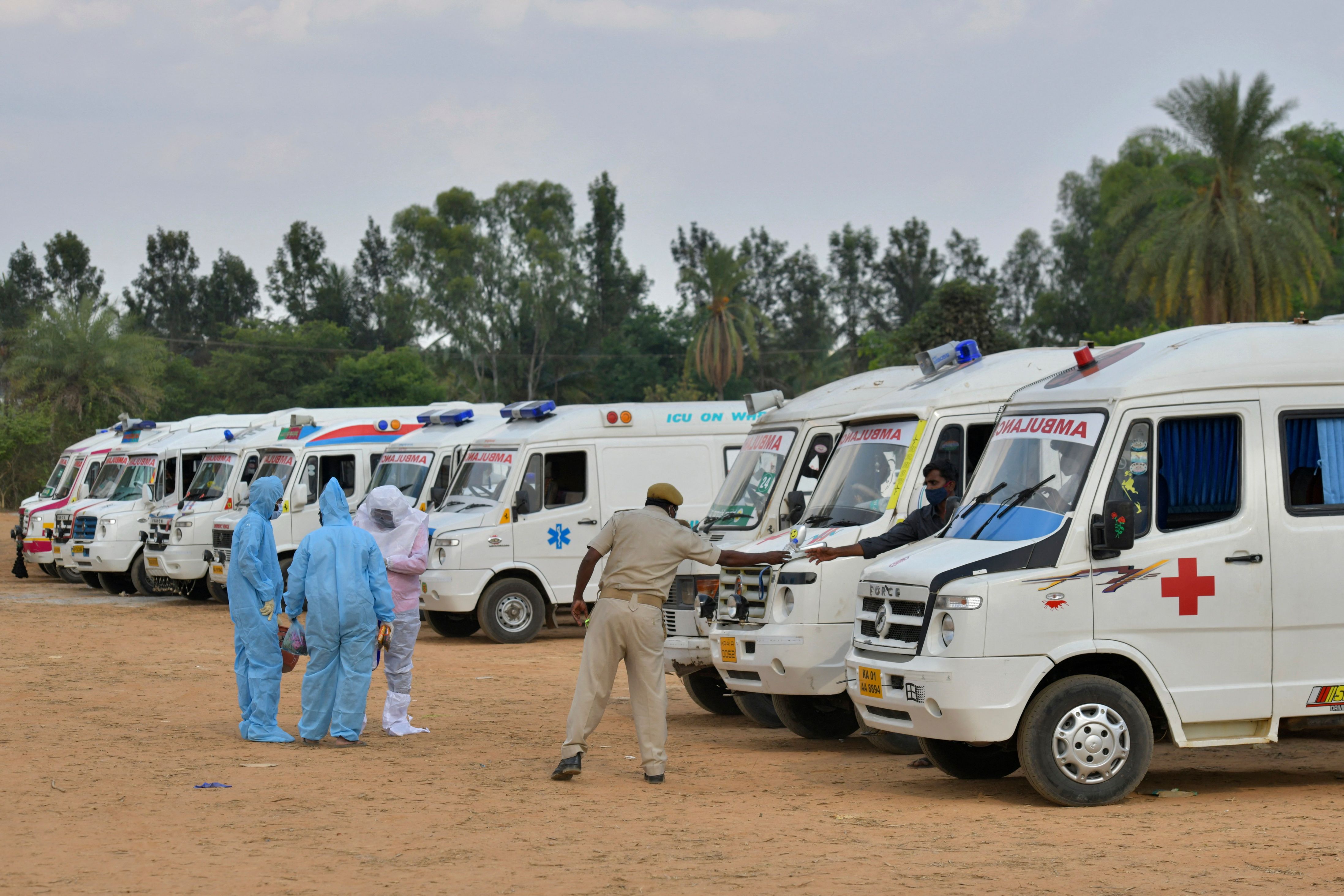 Family members and relatives wearing protective gear stand next to ambulances carrying the bodies of victims who died of the Covid-19 coronavirus at an open-air crematorium set up for the coronavirus victims inside a defunct granite quarry on the outskirts of Bangalore on May 8, 2021. (Photo by Manjunath Kiran / AFP)