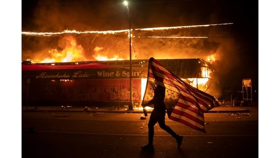 A protester carries a US flag upside down, a sign of distress, next to a burning building, May 28, 2020, in Minneapolis. (Julio Cortez / AP Photo)