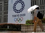 A woman wearing a protective mask walks with an umbrella to shield from the sun walks in front of a Tokyo 2020 display.