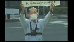 The image shows the man holding a placard in front of the Olympics village.