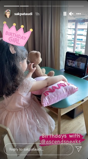 Inaaya in a pink frock along with her teddy bear and customised cushion sat at a table.