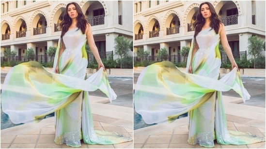 Mahira's gown, decorated with patterned sequins in green, brown, and yellow hues, features a one-shoulder neckline with the other sleeve extending into a flowy train, embellishments on the waist, a bodycon fit, and a floor-sweeping train.