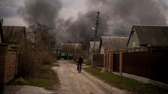 A Ukrainian man rides his bicycle near a factory and a store burning after it had been bombarded in Irpin, on the outskirts of Kyiv, Ukraine.