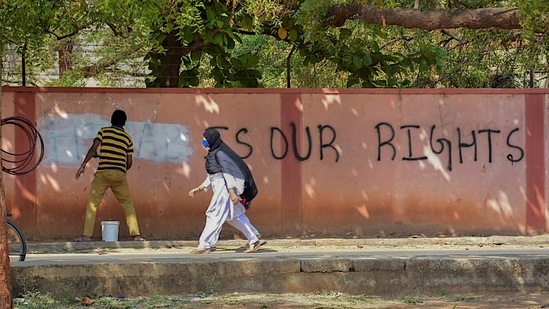 A hijab-wearing student walks past a worker painting a wall to conceal a pro-hijab slogan, Hospet, March 16, 2022 (PTI)