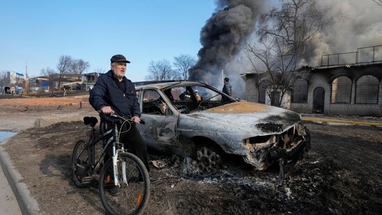 A man rides his bike past the gutted remains of a car and smoke rising from a fire following a Russian attack in Kharkiv, Ukraine(AP)
