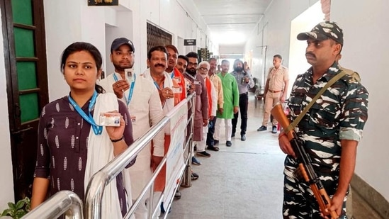 A security person stands guard as electors wait for their turn to cast their vote in the Uttar Pradesh Legislative Council elections. (PTI PHOTO)