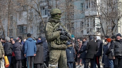 A Russian army soldier stands next to local residents who queue for humanitarian aid delivered during Ukraine-Russia conflict, in the besieged southern port of Mariupol, Ukraine.