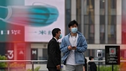 A man wearing a face mask walks by a masked security stands guard at a barricaded entrance displaying a health QR code and a temperature scanner at a commercial office complex, Sunday.