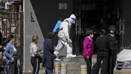 Um trabalhador em um traje de proteção pulveriza desinfetante enquanto moradores usando máscaras fazem fila para testes em massa de coronavírus do lado de fora de um complexo residencial, em Pequim, na terça-feira.  (AP)