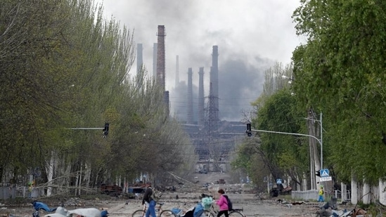 People walk their bikes across the street as smoke rises above a plant of Azovstal Iron and Steel Works during Ukraine-Russia conflict in the southern port city of Mariupol, Ukraine.(REUTERS)