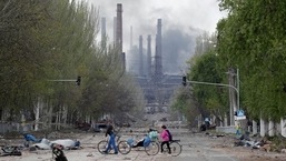 Pessoas andam de bicicleta pela rua enquanto a fumaça sobe acima de uma fábrica da Azovstal Iron and Steel Works durante o conflito Ucrânia-Rússia em Mariupol, 