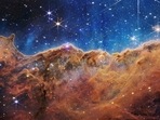 The ‘Cosmic Cliffs' of the Carina Nebula is seen in an image divided horizontally by an undulating line between a cloudscape forming a nebula along the bottom portion and a comparatively clear upper portion.