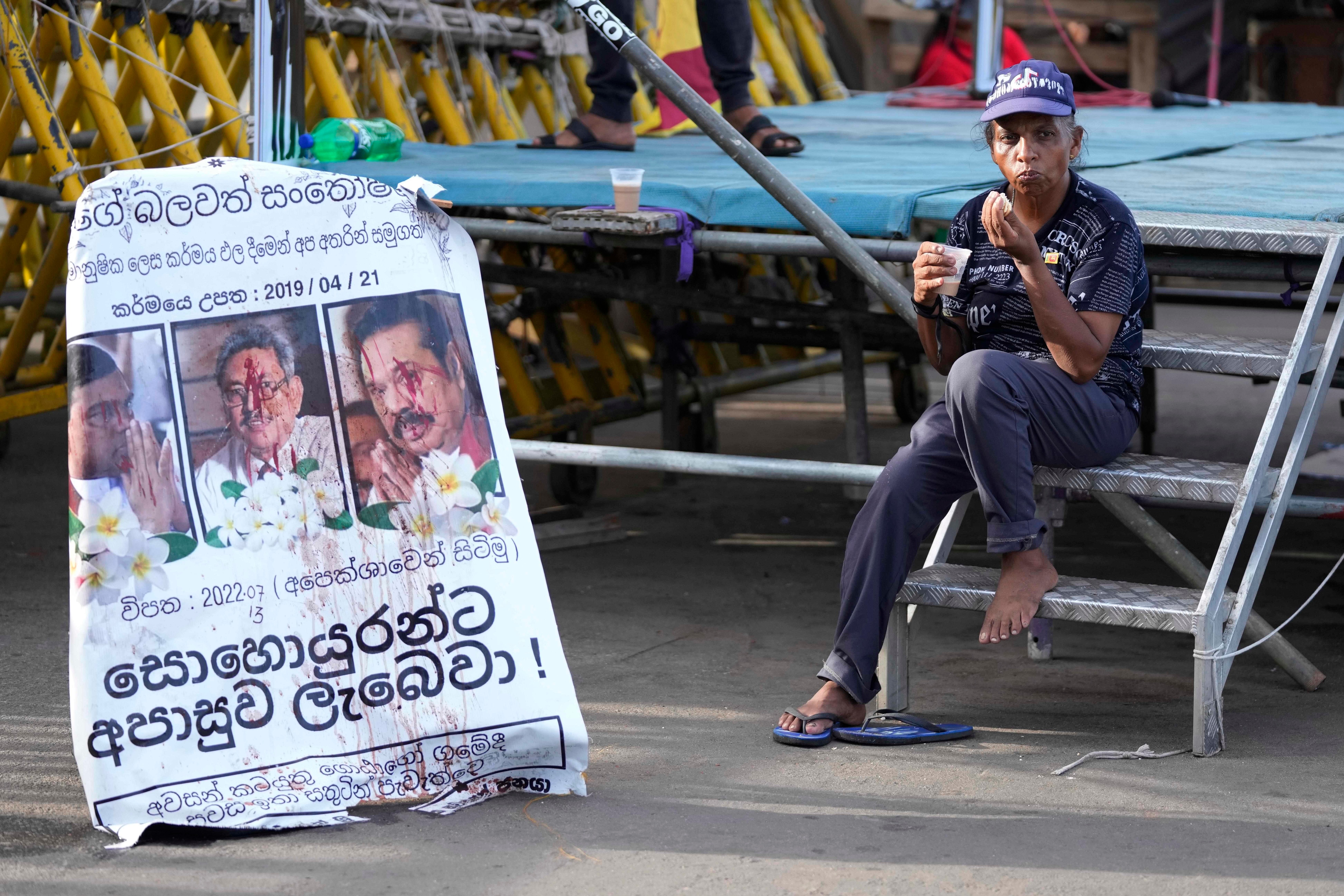 A protester drinks a cup of tea as she sits by a defaced poster carrying portraits of ousted president Gotabaya Rajapaksa, center, and his brothers at the entrance to president's office in Colombo, Sri Lanka on Friday.(AP) A protester drinks a cup of tea as she sits by a defaced poster carrying portraits of ousted president Gotabaya Rajapaksa, center, and his brothers at the entrance to president's office in Colombo, Sri Lanka on Friday.(AP)