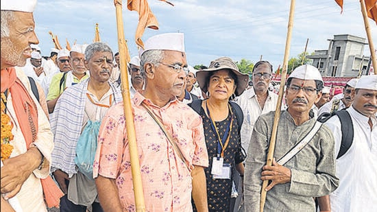 Nandini Dias walking with the flag bearers leading the Yatra, followed by chanters, dholak players and cymbal players.