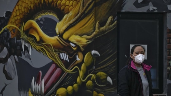 Covid In China: A woman wearing a face mask stands near a mural depicting a dragon in Beijing. Covid In China: A woman wearing a face mask stands near a mural depicting a dragon in Beijing.