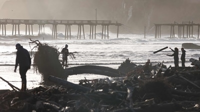 People gather near storm debris washed up on the beach, with a damaged pier in the background, on January 10 in Capitola, California. Storms have toppled trees, flooded roads and cut power to tens of thousands of people. The Pacific storms are expected to bring more rain and wind through the end of the week, as reported by AFP. ( Mario Tama / Getty Images via AFP)