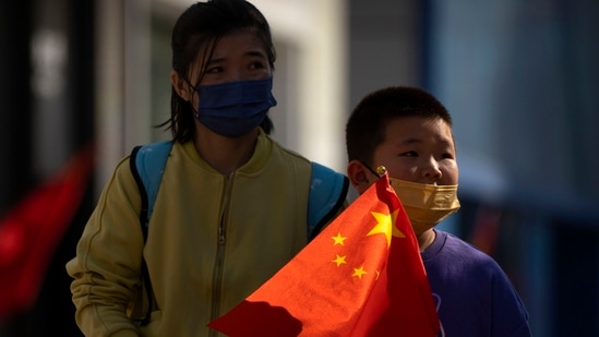 China Population Policy: A boy wearing a face mask carries a Chinese flag as he walks along a pedestrian shopping street.(AP)