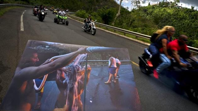 A printed photograph taken on September 28, 2017 shows people bathing on the highway after Hurricane Maria, as motorcyclists ride past the same location on May 27, 2018. FEMA officials said that longstanding rules prevent federal officials from granting reconstruction aid to victims who don’t have titles to homes. In many cases, storm victims were informally given property by a relative or inherited without a will or deed. (Ramon Espinosa / AP)