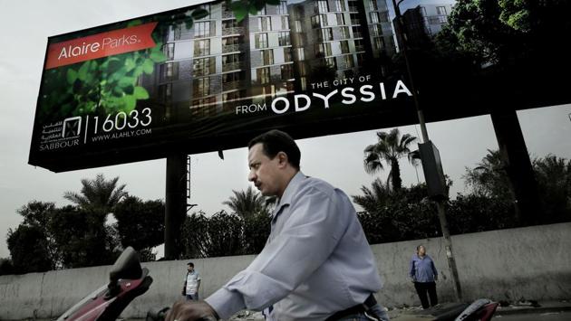 A man rides his motorbike as others wait for a bus under a billboard promoting new residential housing, in Cairo, Egypt. Billboards across Cairo advertise luxury homes with “breathtaking” views and names like “La Verde” or “Vinci” in Egypt’s new capital that is under construction in the desert, miles from the Nile-side city which has been the seat of power for more than 1,000 years. (Nariman El-Mofty / AP) A man rides his motorbike as others wait for a bus under a billboard promoting new residential housing, in Cairo, Egypt. Billboards across Cairo advertise luxury homes with “breathtaking” views and names like “La Verde” or “Vinci” in Egypt’s new capital that is under construction in the desert, miles from the Nile-side city which has been the seat of power for more than 1,000 years. (Nariman El-Mofty / AP)