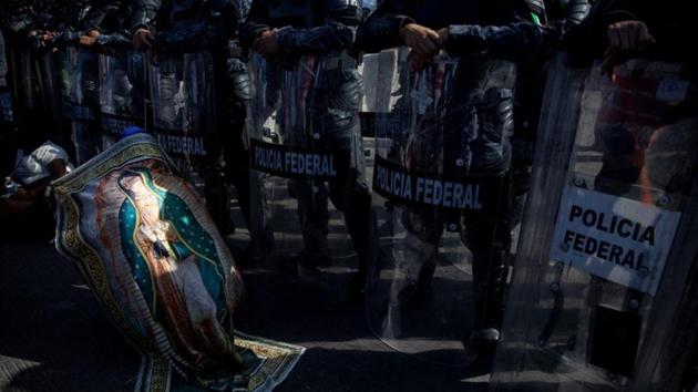 On their way to the US border, the migrants walked the length of Mexico. Here, the Virgin of Guadalupe - an image of the Virgin Mary who devotees believe appeared to an indigenous man in the 16th century -- looms large. As if seeking her protection, a man draped himself with a banner depicting her as he crouched before a phalanx of riot police in Tijuana. (Alkis Konstantinidis / Reuters) On their way to the US border, the migrants walked the length of Mexico. Here, the Virgin of Guadalupe - an image of the Virgin Mary who devotees believe appeared to an indigenous man in the 16th century -- looms large. As if seeking her protection, a man draped himself with a banner depicting her as he crouched before a phalanx of riot police in Tijuana. (Alkis Konstantinidis / Reuters)
