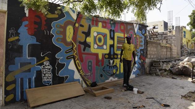 One mural features a fist raised against a hypnotic-blue background, for example. Underneath it, carpenters work steadily at wood pallets. Another mural has two mysterious women set against brilliant indigos and azures staring into the distance. A real-life cow rummages on the ground at their feet. (Seyllou / AFP)