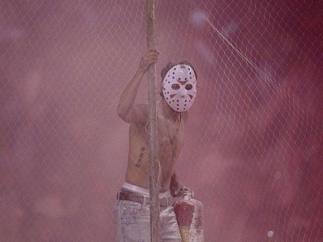 A fan of Ecuador's Liga Deportiva Universitaria soccer team wears a mask as he watches from behind a fence a Copa Sudamericana soccer game against Paraguay's Libertad in Quito, Ecuador.