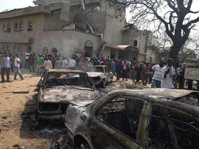 Onlookers gather around a destroyed car at the site of a bomb blast, which killed atleast 25, at St Theresa Catholic Church in Madalla, Nigeria. AP Photo
