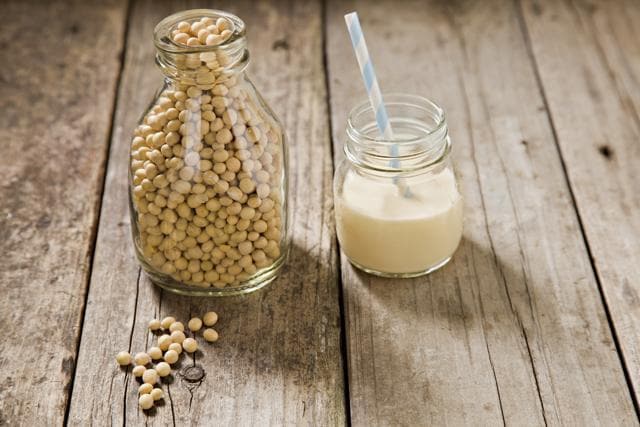A close up shot of an old milk bottle filled with dry soy beans with a few beans on the foreground and a small glass jar container with soy milk and a straw. Shot on an old grungy wooden table. (Getty Images)
