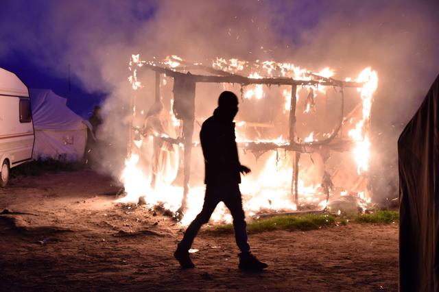 A migrant walks past a shack set on fire during the demolition of the " camp. (AFP) A migrant walks past a shack set on fire during the demolition of the " camp. (AFP)