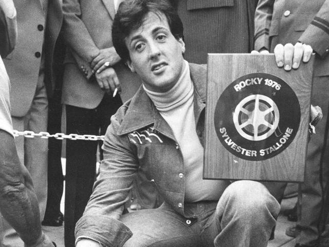 In this September 22, 1977 photo, actor and screenwriter Sylvester Stallone poses with a terrazzo inlay to be placed in the streets of the Westwood section of Los Angeles. He had become a big star after Rocky, which was released on November 21, 1976. (AP Photo)