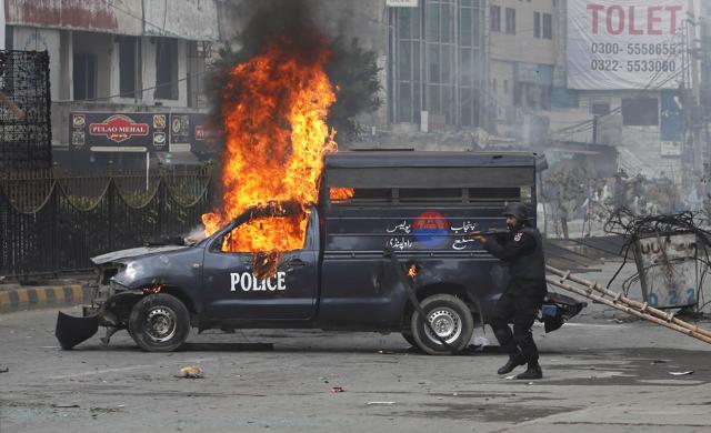 A Pakistani police officer aims his gun towards the protesters next to a burning police vehicle during a clash in Islamabad on November 25, 2017. (AP) A Pakistani police officer aims his gun towards the protesters next to a burning police vehicle during a clash in Islamabad on November 25, 2017. (AP)