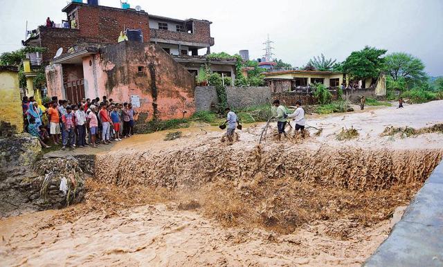 People crossing the overflowing Patiala-ki-Rao rivulet amid rain near Singha Devi temple at Nayagaon on the outskirts of Chandigarh on Monday. (HT Photo)