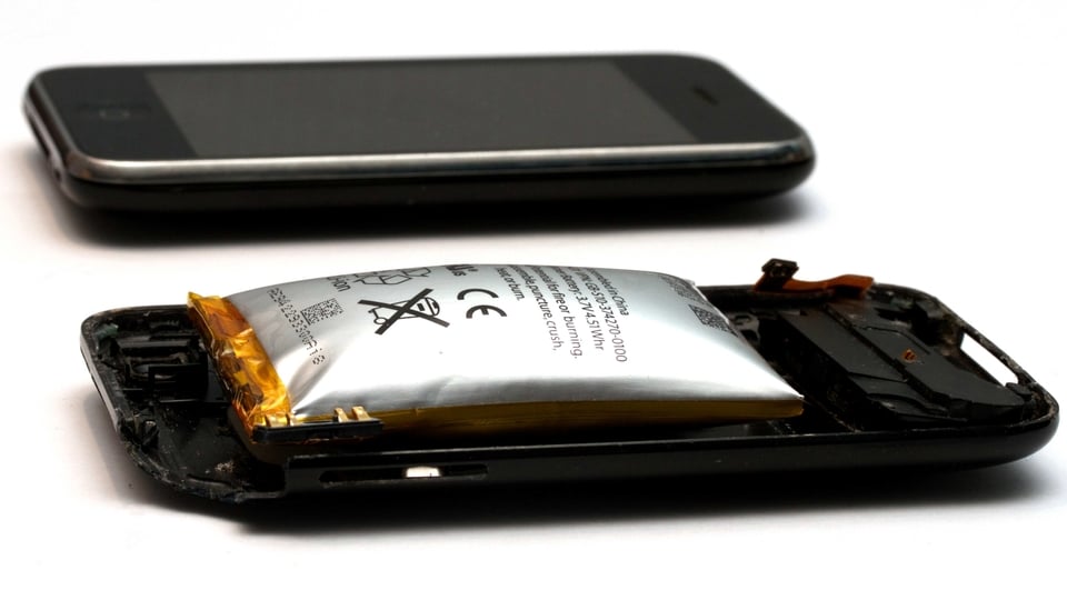An Apple iPhone 3GS's Lithium-ion polymer battery, which has expanded due to a short circuit failure. The battery is shown in situ on top of the rear phone case; pictured behind is an intact iPhone 3GS for size comparison.